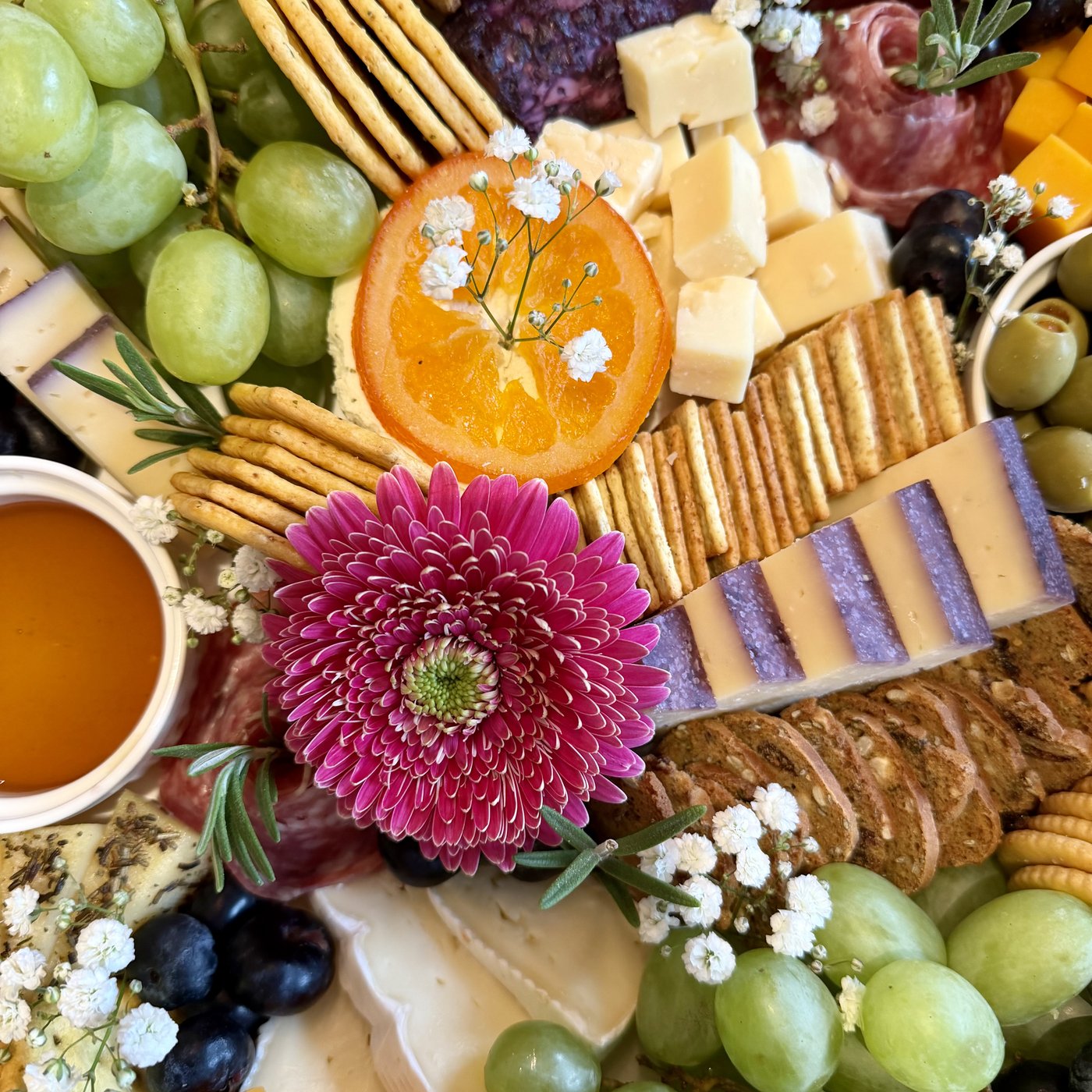Close-up of a board with a pink gerbera daisy, a candied orange slice, green grapes, cheeses, crackers and baby's breath