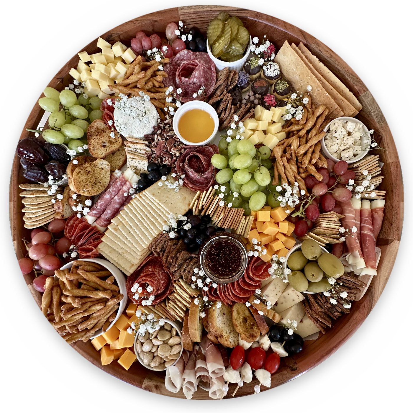 A round wooden board filled with cheeses, cured meats, grapes, crackers, olives, honey and baby's breath