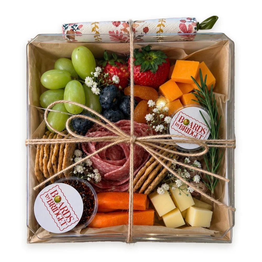 Snack box tied with twine and a floral napkin: grapes, strawberries, blueberries, cheddar, a salami rose, crackers, carrots, cheese cubes and fig jam